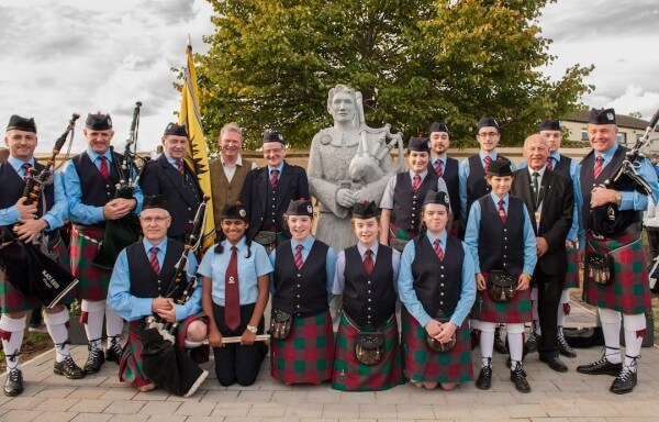 Pipe band in kilts and dark jackets posed in two rows around the stone Thomas Ashe memorial statue at its unveiling.